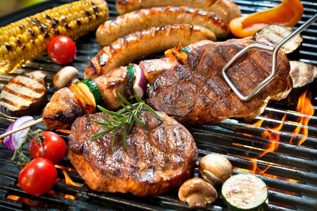 Close-up of a sizzling BBQ grill loaded with grilled steaks, sausages, vegetable skewers, corn on the cob, and cherry tomatoes, creating a vibrant and flavorful outdoor cooking scene.